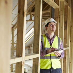 man in hard hat and safety vest inspects house framing with clipboard