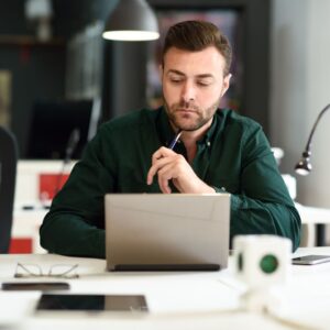 man in green shirt sits in front of laptop holding a pen
